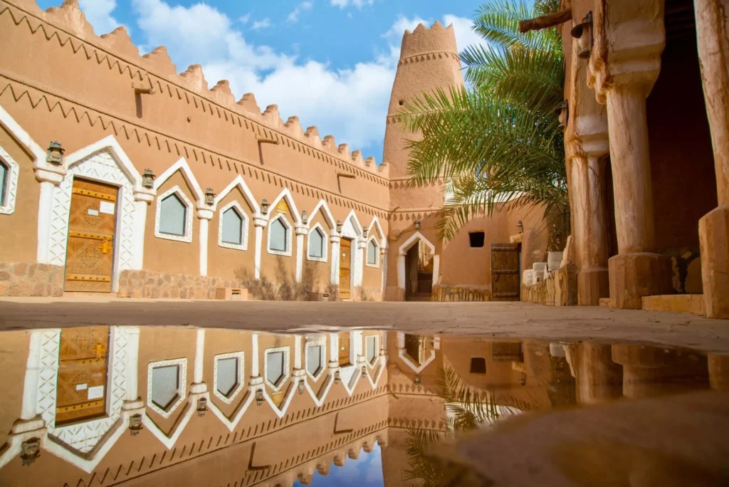 A traditional mud-brick building with an open courtyard, showing reflections of the architecture in a puddle after rain.
