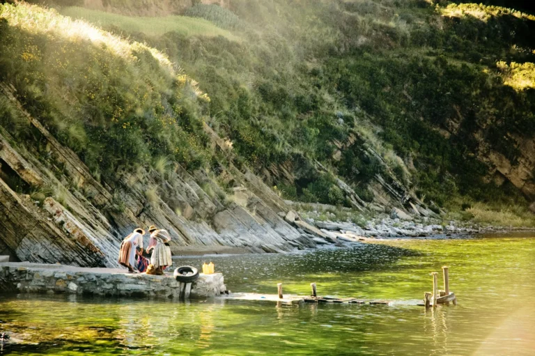 A serene natural pool with clear green water, surrounded by lush vegetation, and people relaxing by the water's edge.