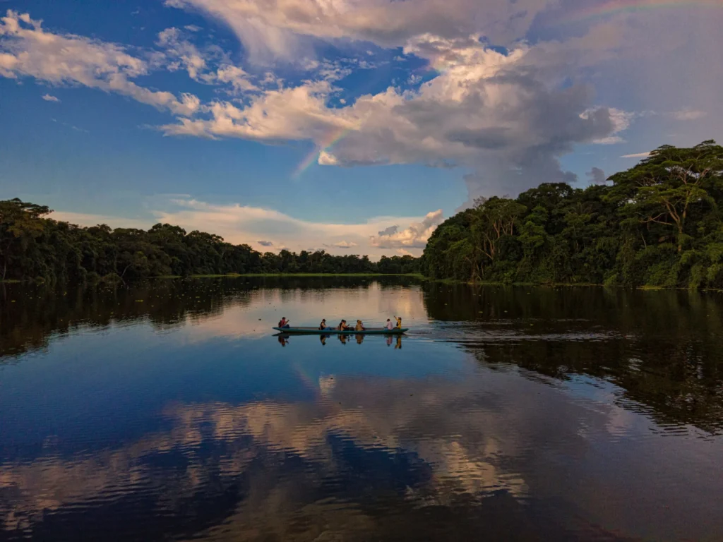 A serene river or lake reflecting the sky and surrounding trees, under a wide sky with scattered clouds, likely in the Amazon rainforest.