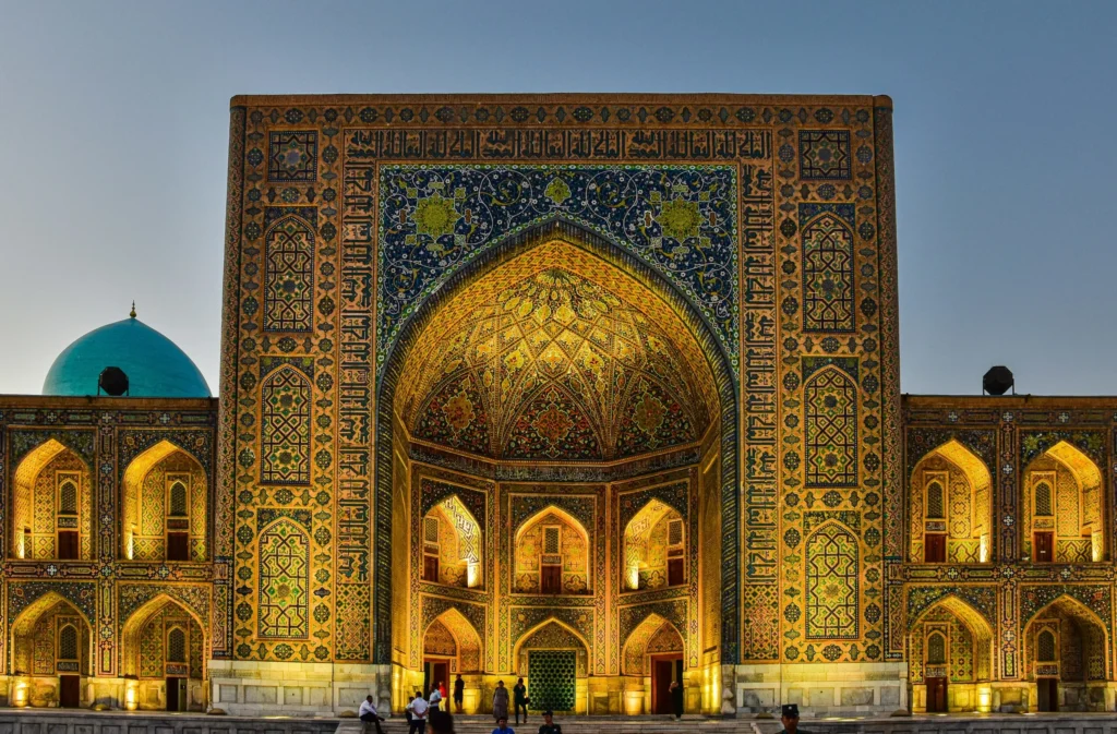 An ornate, ancient madrasah (Islamic school) with intricate tilework and a large arched entrance, illuminated at dusk.