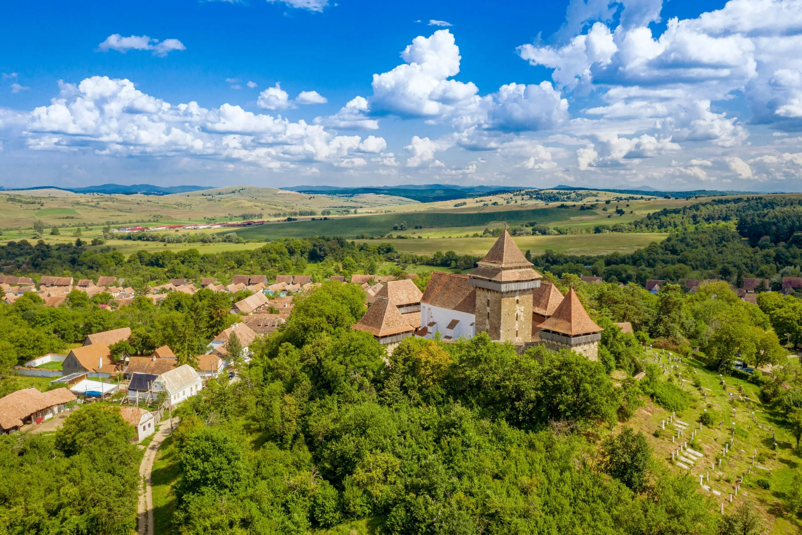 An aerial view of a fortified church or castle nestled within a green, hilly landscape with a village nearby.