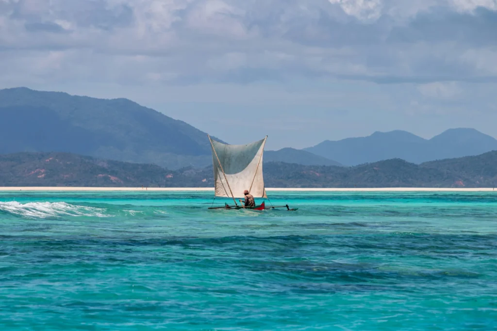 A traditional sailing boat with a white sail glides across turquoise water with mountains in the background.
