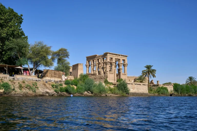 A view of the Philae Temple complex across a body of water, with lush greenery and a clear blue sky.