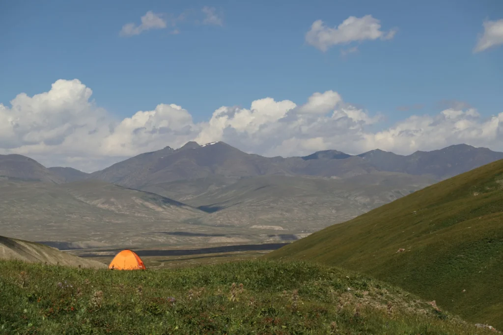 A vast, open landscape with rolling hills and a single orange tent pitched in the middle ground, under a partly cloudy sky.