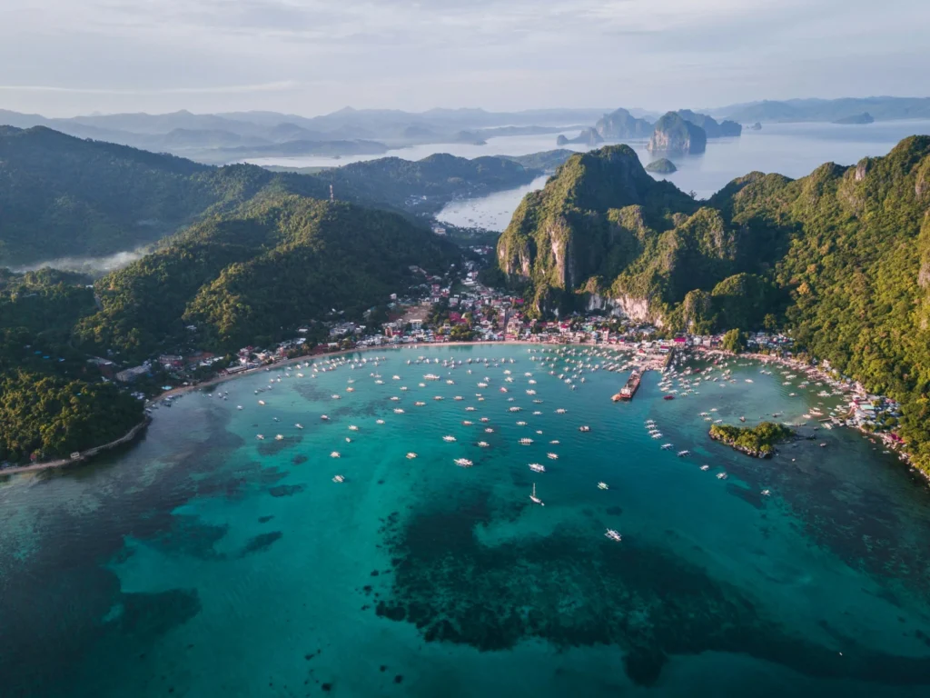 An aerial view of a picturesque bay with a village nestled among lush green hills and clear blue water filled with boats.