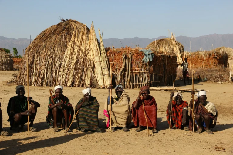 A group of people in traditional attire are seated in front of thatched huts.
