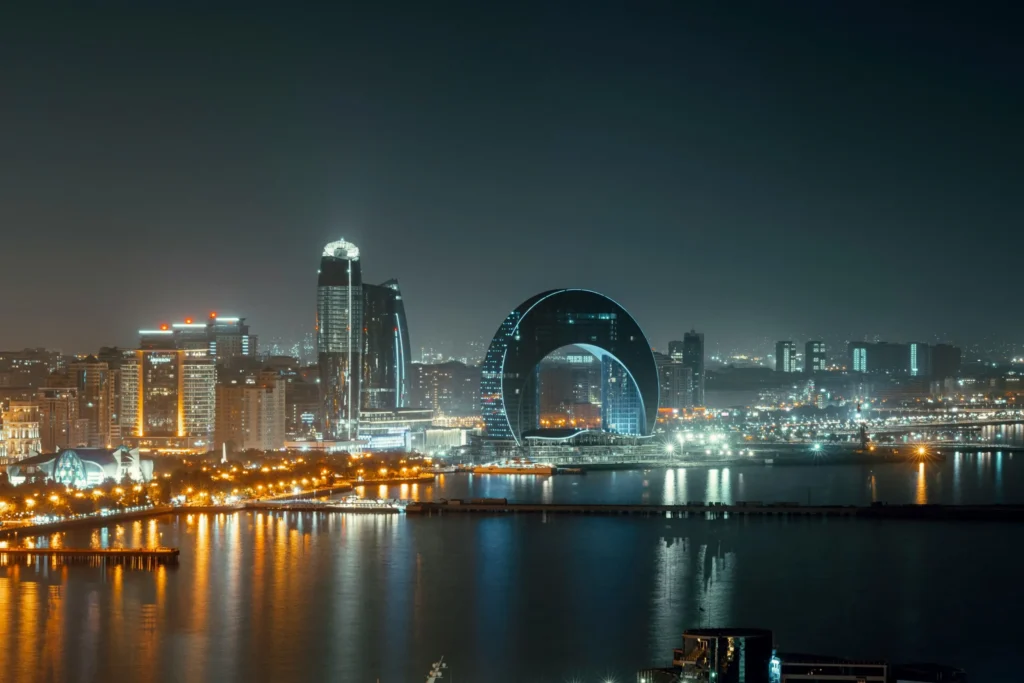 A cityscape at night with illuminated skyscrapers and a distinctive, ring-shaped building reflected in the water.