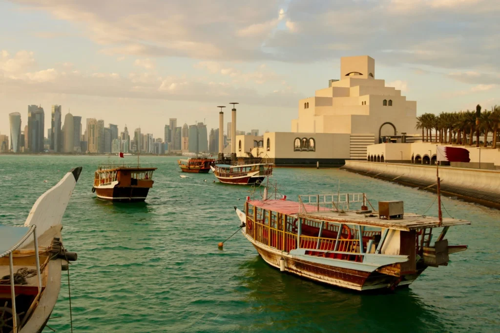 Traditional wooden boats (dhows) on the water with a modern city skyline and a distinctive white museum building in the background.