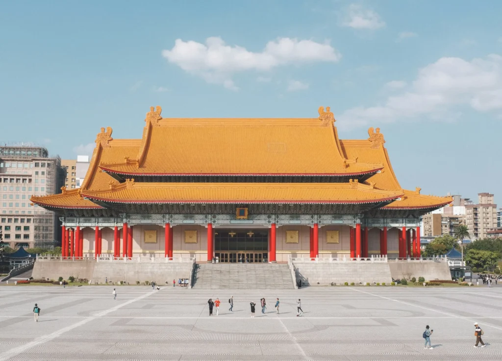 A wide view of a large, traditional Chinese-style building with a red roof and ornate details, set in a spacious plaza under a blue sky.