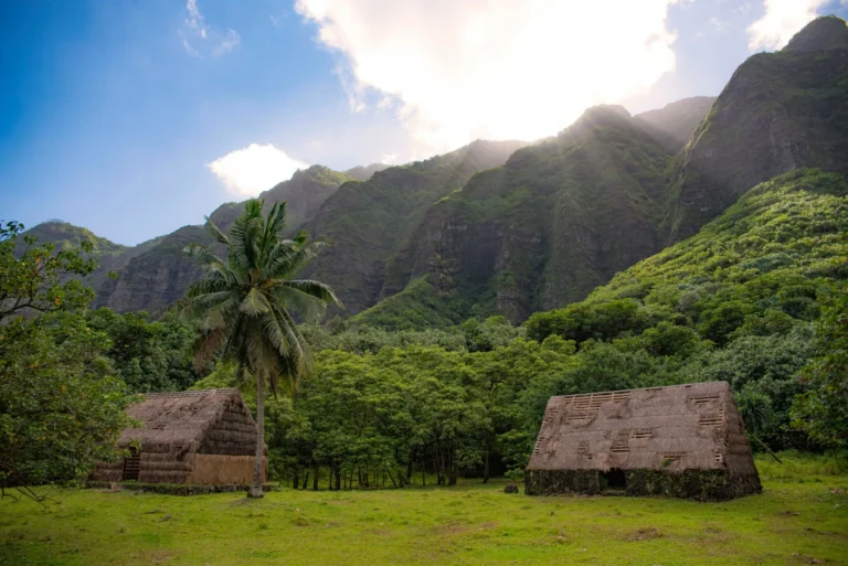 Two traditional huts are nestled in a lush green valley with towering mountains in the background.