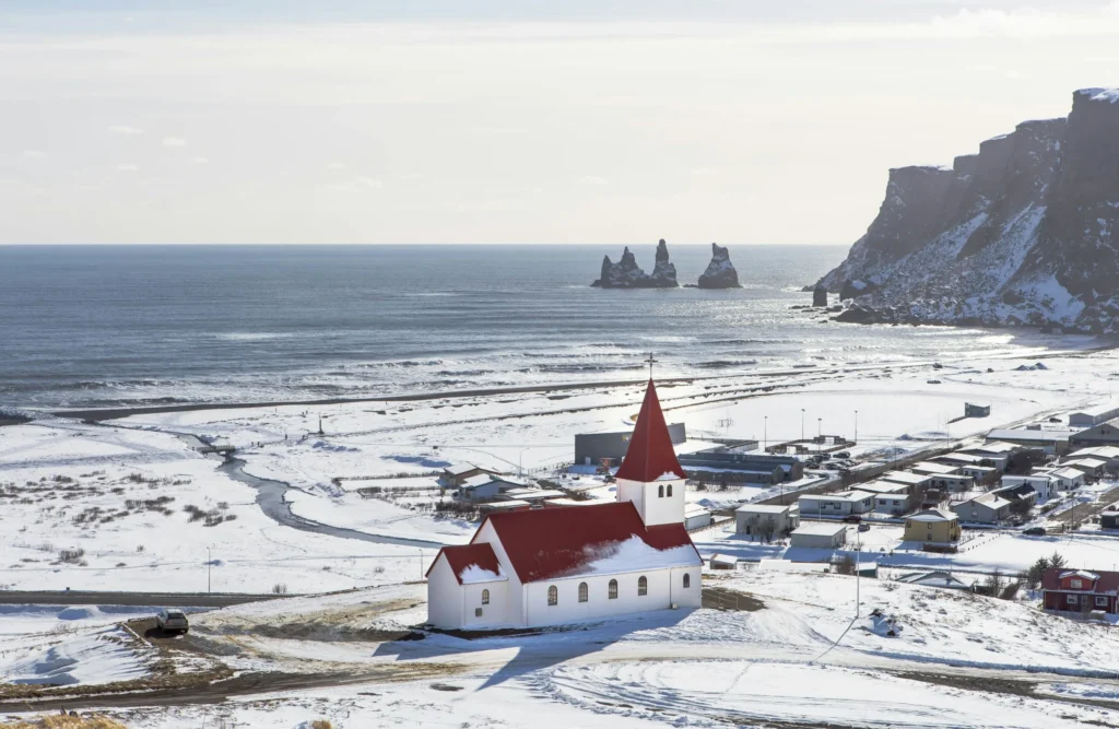 A snow-covered coastal landscape with a red-roofed building and a lighthouse.