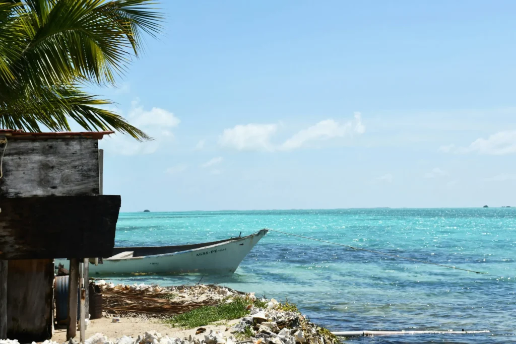 A rustic wooden structure on a small patch of land next to crystal clear turquoise water, with a small boat nearby and palm trees.