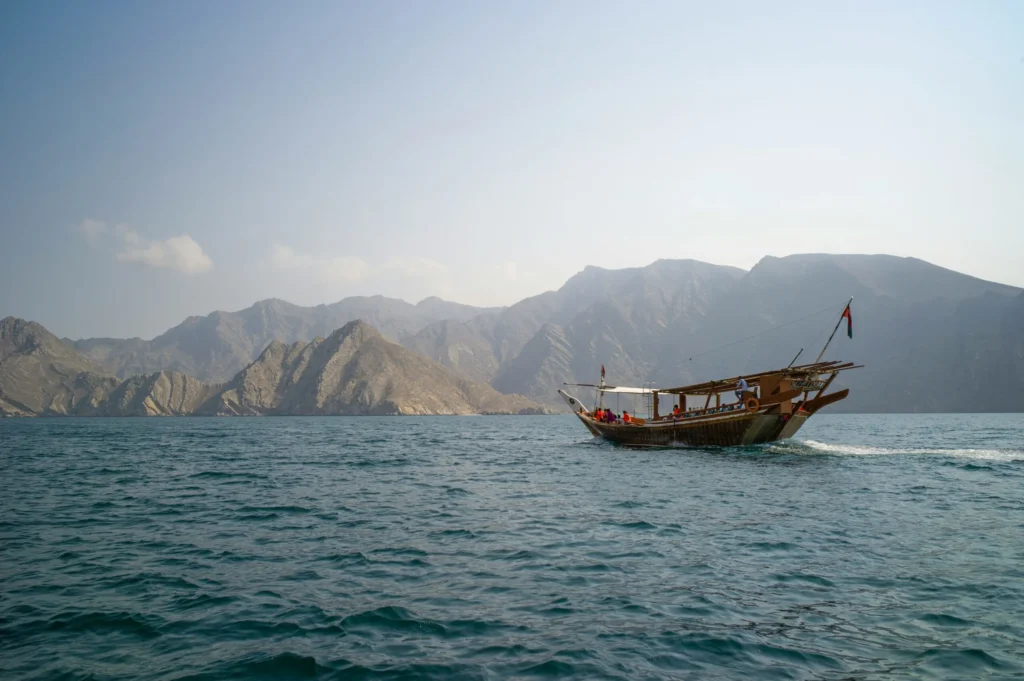 A traditional wooden boat (dhow) sails on the sea with rugged, mountainous coastline in the background.