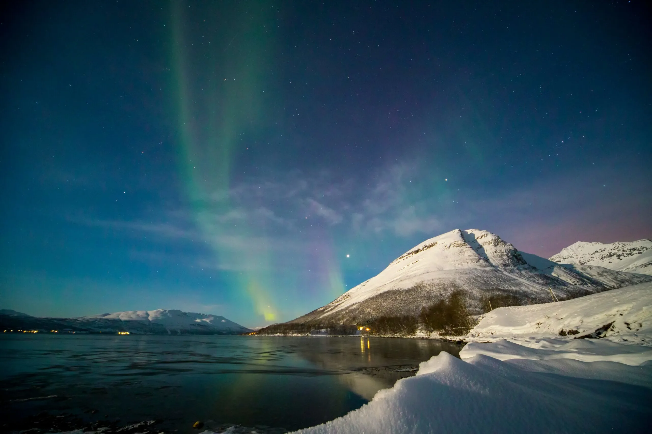 The Aurora Borealis, or Northern Lights, illuminating the sky above a snowy mountain and a body of water.