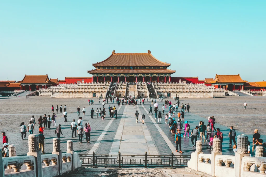 A wide view of the Forbidden City with its traditional red and yellow buildings, and a large courtyard filled with people.