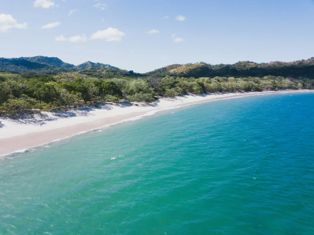 An aerial view of a wide, curving beach with turquoise water and green hills in the background.