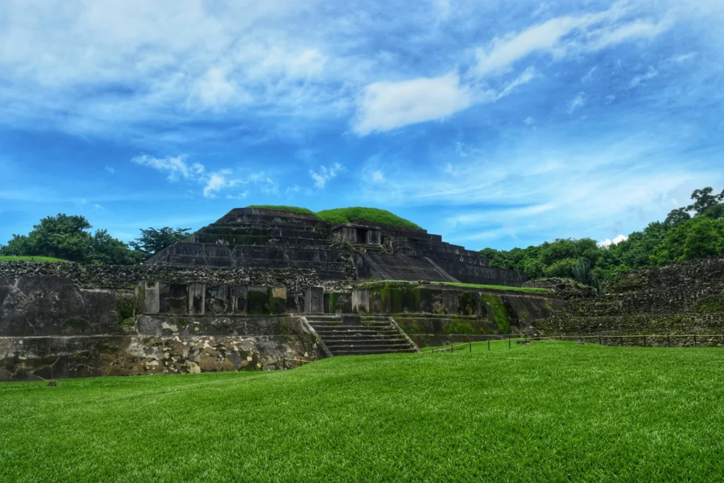 Ancient ruins made of stone blocks, partially covered by grass, under a vast blue sky with scattered clouds.
