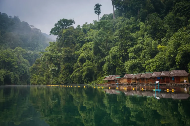A serene lake with wooden stilt houses on the shore, surrounded by lush green mountains under a misty sky.