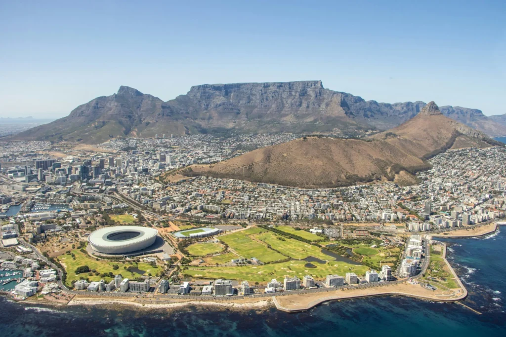 An aerial view of Cape Town, South Africa, with Table Mountain in the background, showing the city, stadium, and coastline.