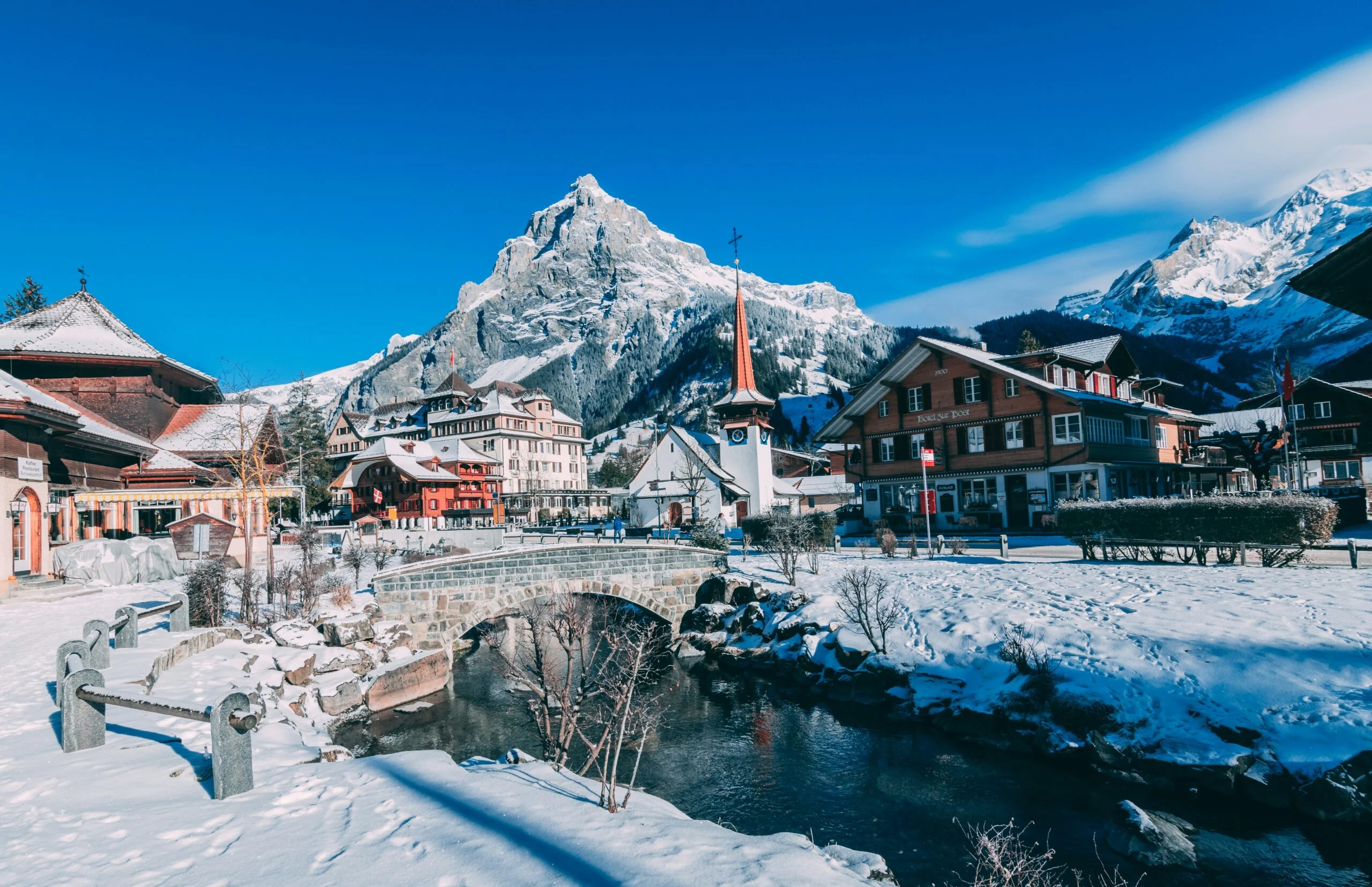 A snow-covered village nestled beside a frozen lake with dramatic mountains in the background.