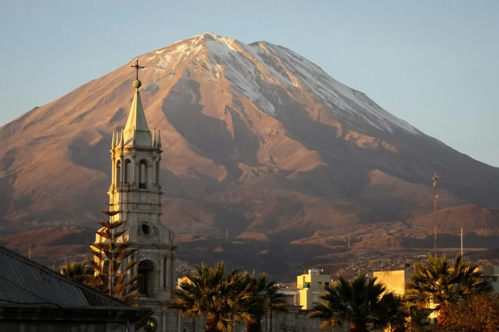 A grand cathedral with a tall spire in the foreground, set against the backdrop of a large, conical volcano.