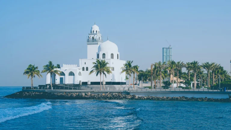 A white mosque with a single dome and minaret, situated on a small island or peninsula surrounded by water.