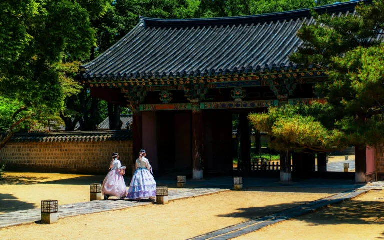 A traditional Korean temple gate with two figures in traditional dress approaching it, surrounded by lush trees.