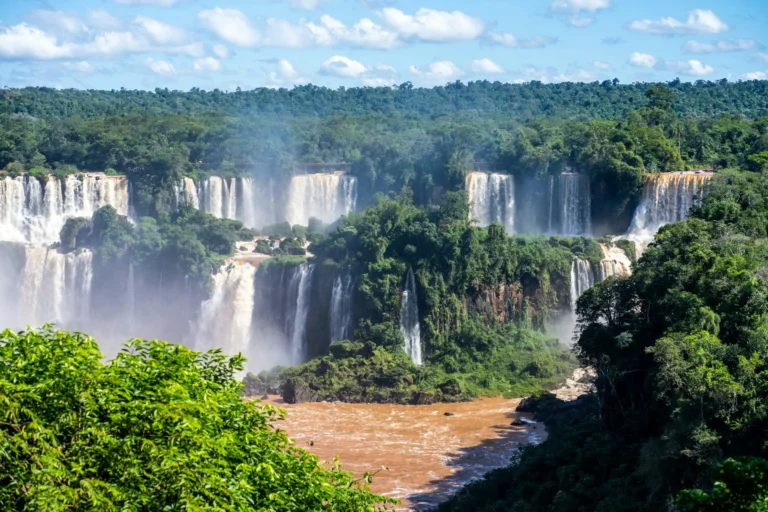 A wide shot of Iguazu Falls, showing multiple cascades surrounded by lush green forest.
