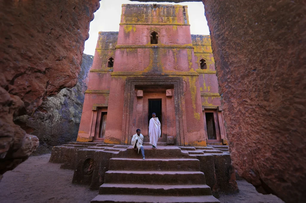 An ancient rock-hewn church with reddish-brown walls and intricate carvings, with a person standing at the entrance.