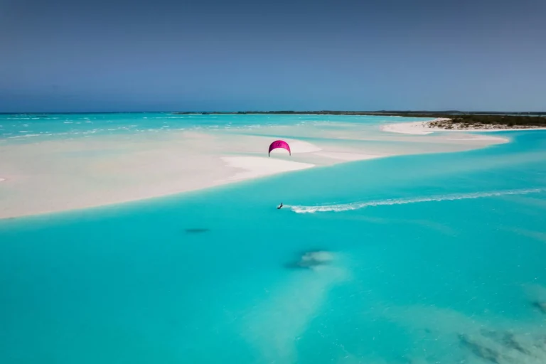 An aerial view of a pristine sandbar surrounded by clear turquoise water, with a kite surfer.