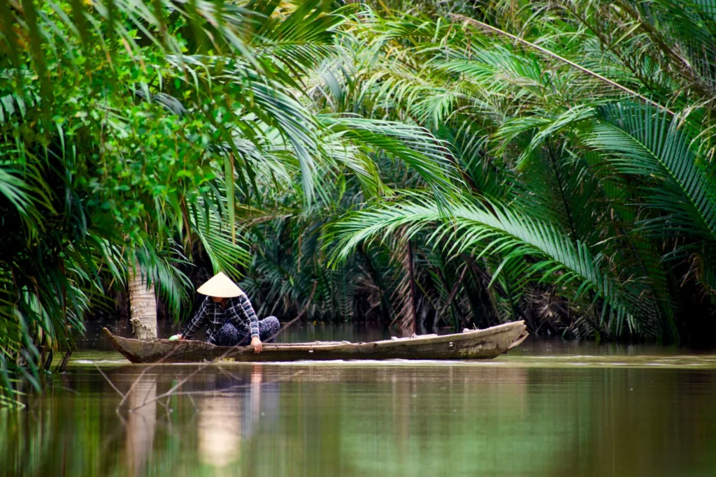 A traditional wooden boat with two people in conical hats navigating a calm river surrounded by dense tropical foliage.