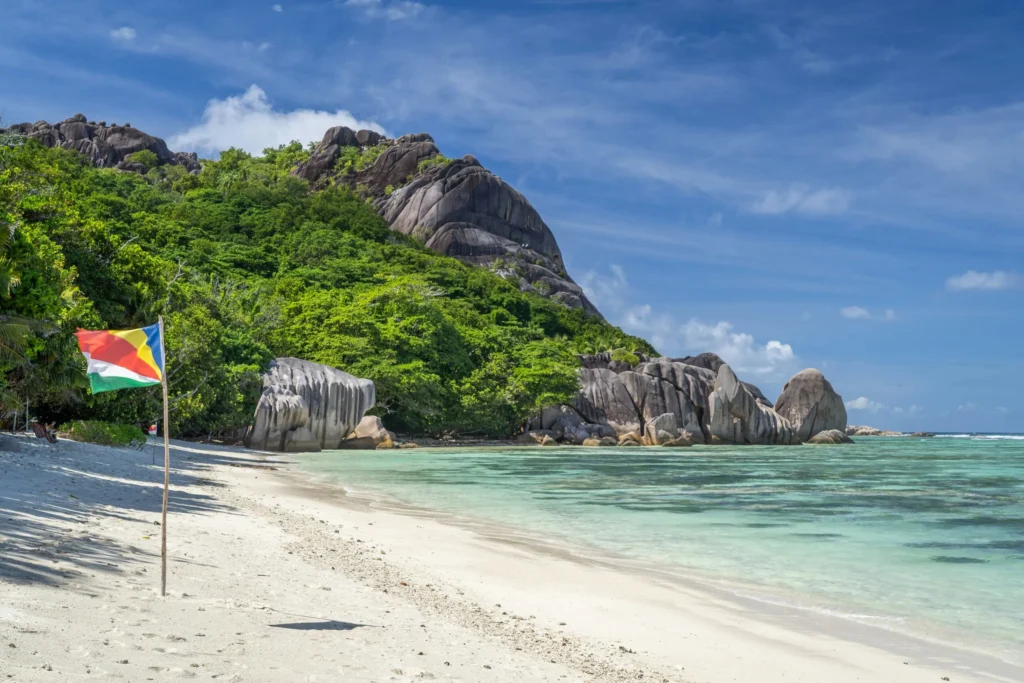 A white sand beach with granite rock formations and a flag, under a blue sky with clouds.