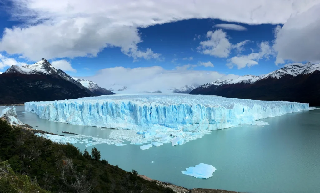 A vast glacier (Perito Moreno Glacier) with a large lake in front of it, surrounded by mountains under a partly cloudy sky.