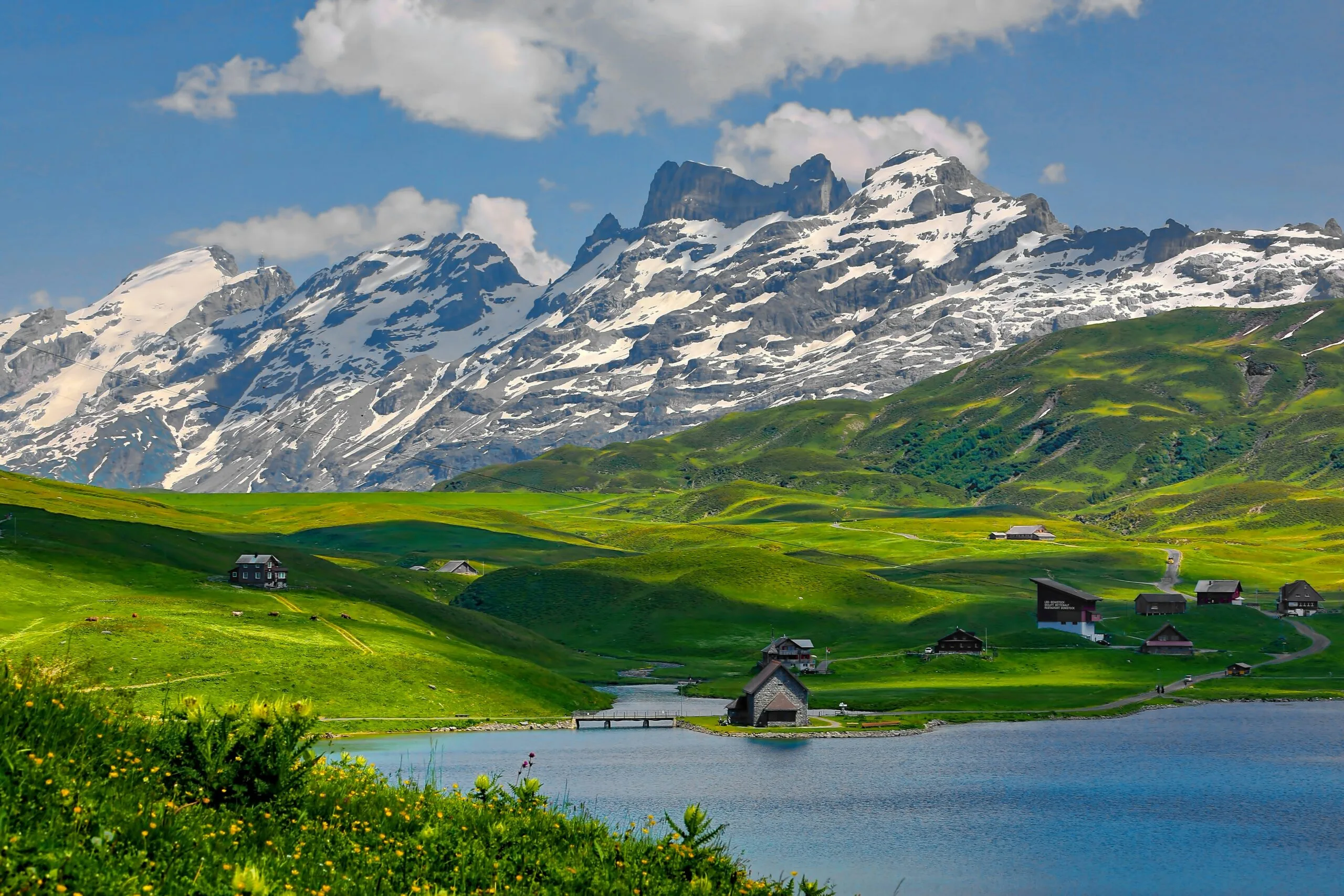 A serene mountain lake surrounded by green hills and distant snow-capped peaks.