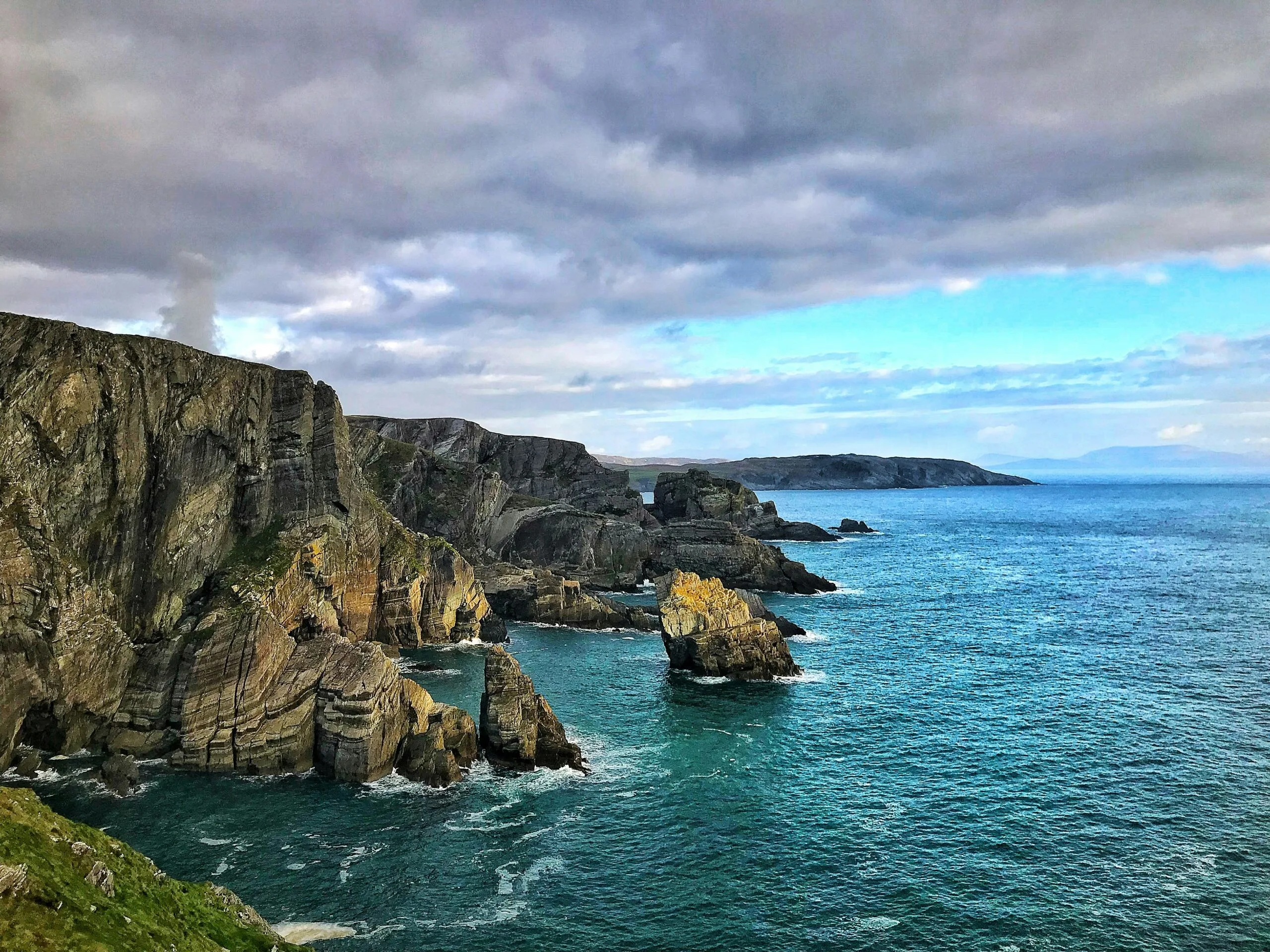 Rugged sea cliffs with rock formations jutting into the blue ocean under a cloudy sky.