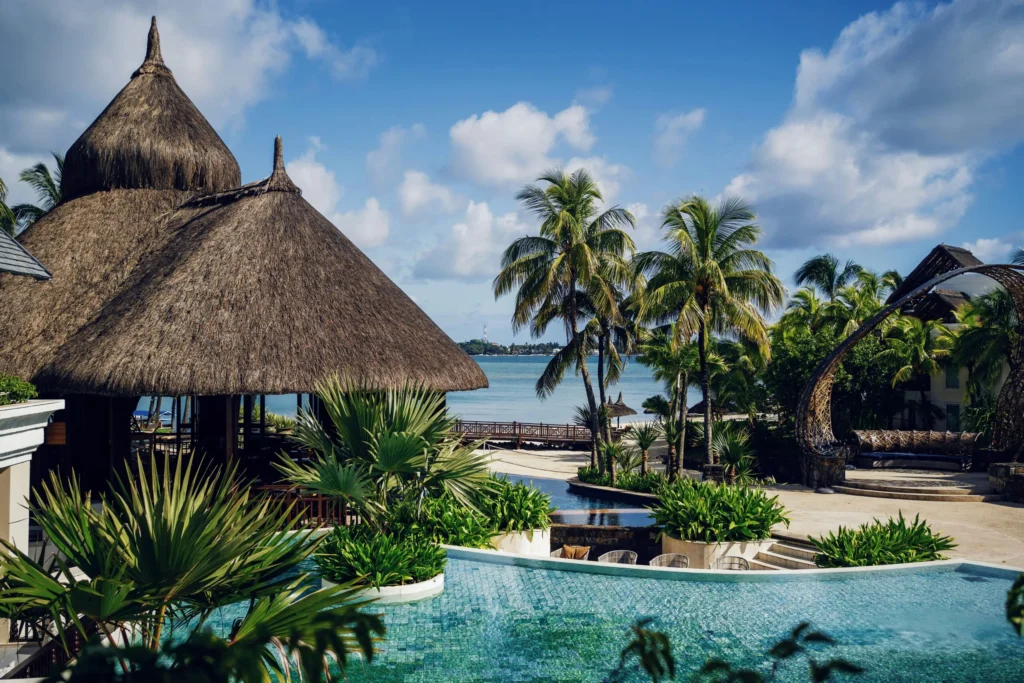 A resort area with thatched-roof buildings, swimming pools, and palm trees, overlooking a body of water.