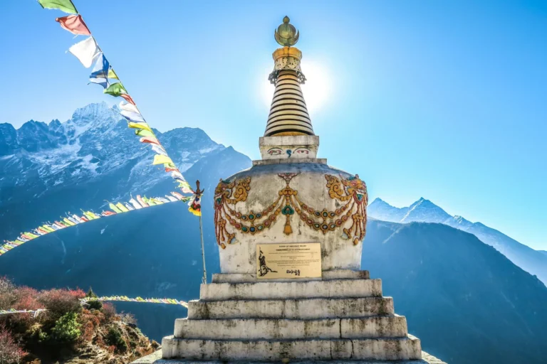 A traditional white stupa with prayer flags, set against a backdrop of snow-capped mountains under a bright blue sky.