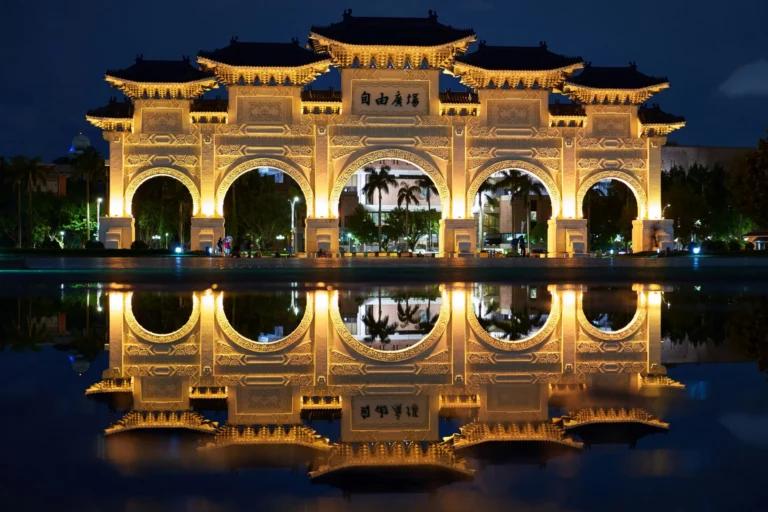A grand, ornate traditional Chinese gate illuminated at night, with its reflection in water.