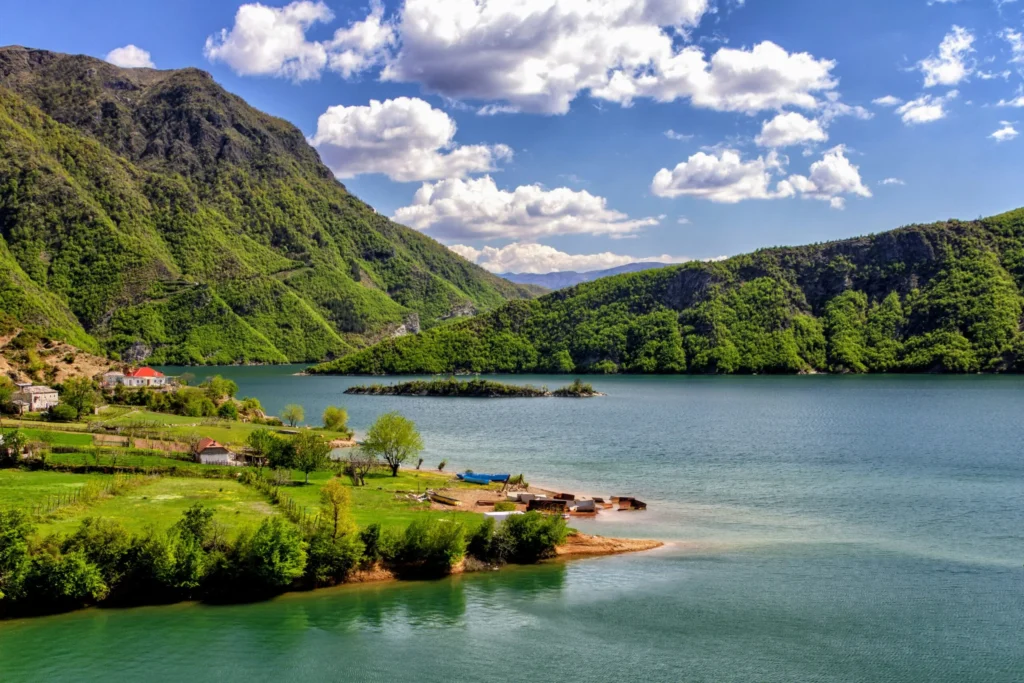 A picturesque lake surrounded by green hills with a small village on the shore under a partly cloudy sky.