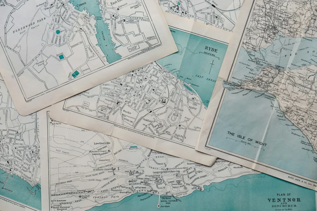 A collection of vintage-style maps of the Isle of Wight, are spread out, with some overlapping, showing coastal areas and town plans.