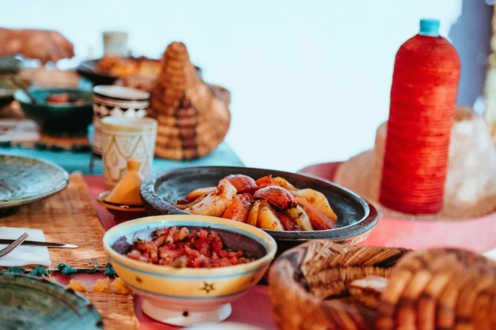 A close-up of a table set with traditional Moroccan dishes, including a tagine-like dish with vegetables, a bowl of salsa or chopped salad, and various woven baskets and ceramic cups. A red bottle is visible in the background.
