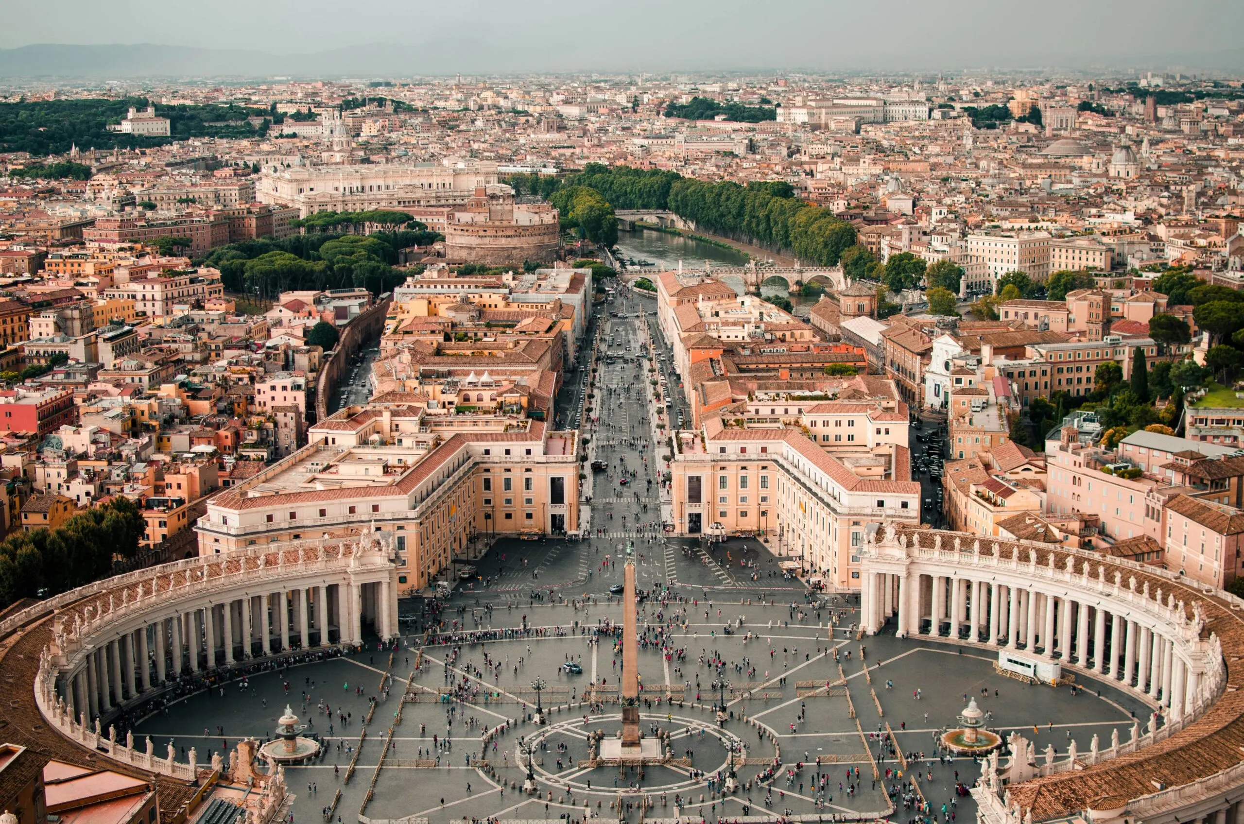 An aerial or high-angle view of St. Peter's Square, showing its distinctive elliptical shape and the obelisk at its center, with the city stretching into the background.
