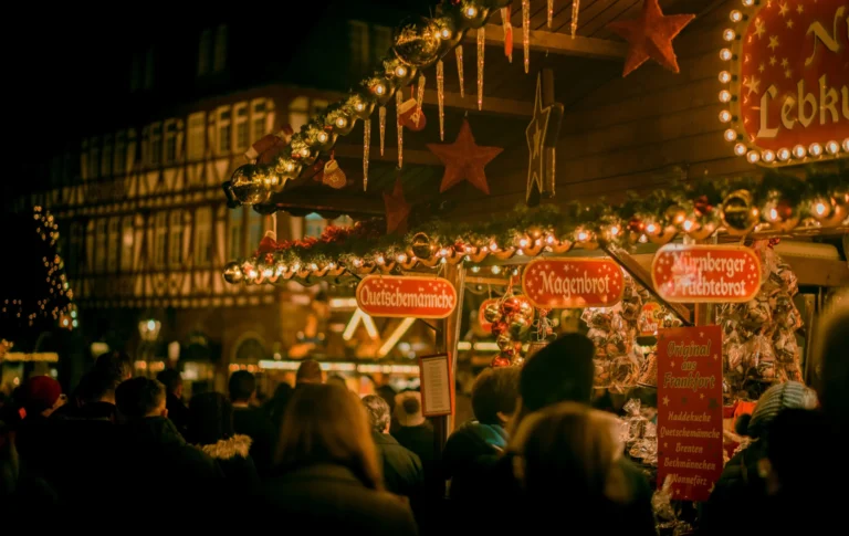 A bustling outdoor night Christmas market, with numerous stalls illuminated by warm lights and a crowd of people.