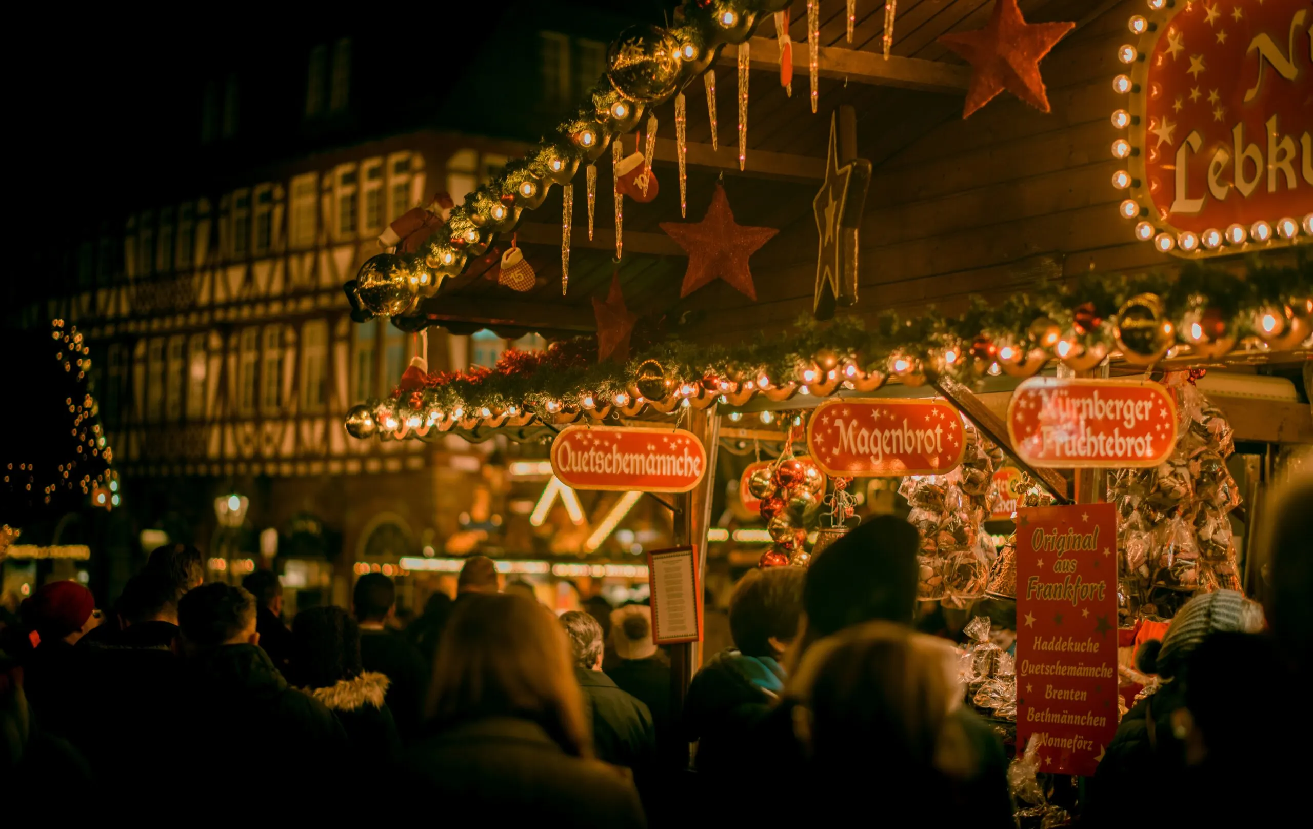 A bustling outdoor night Christmas market, with numerous stalls illuminated by warm lights and a crowd of people.