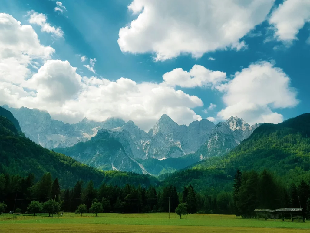 A serene clear turquoise river or lake with a pebbled shoreline, framed by majestic green and brown mountains under a bright sky.