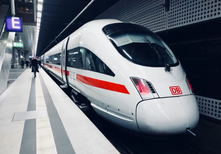 A sleek white high-speed train, a German ICE, with a red stripe and DB logo, is stopped at a modern underground station platform. A person walks away in the distance, and a blue "E" sign is visible.