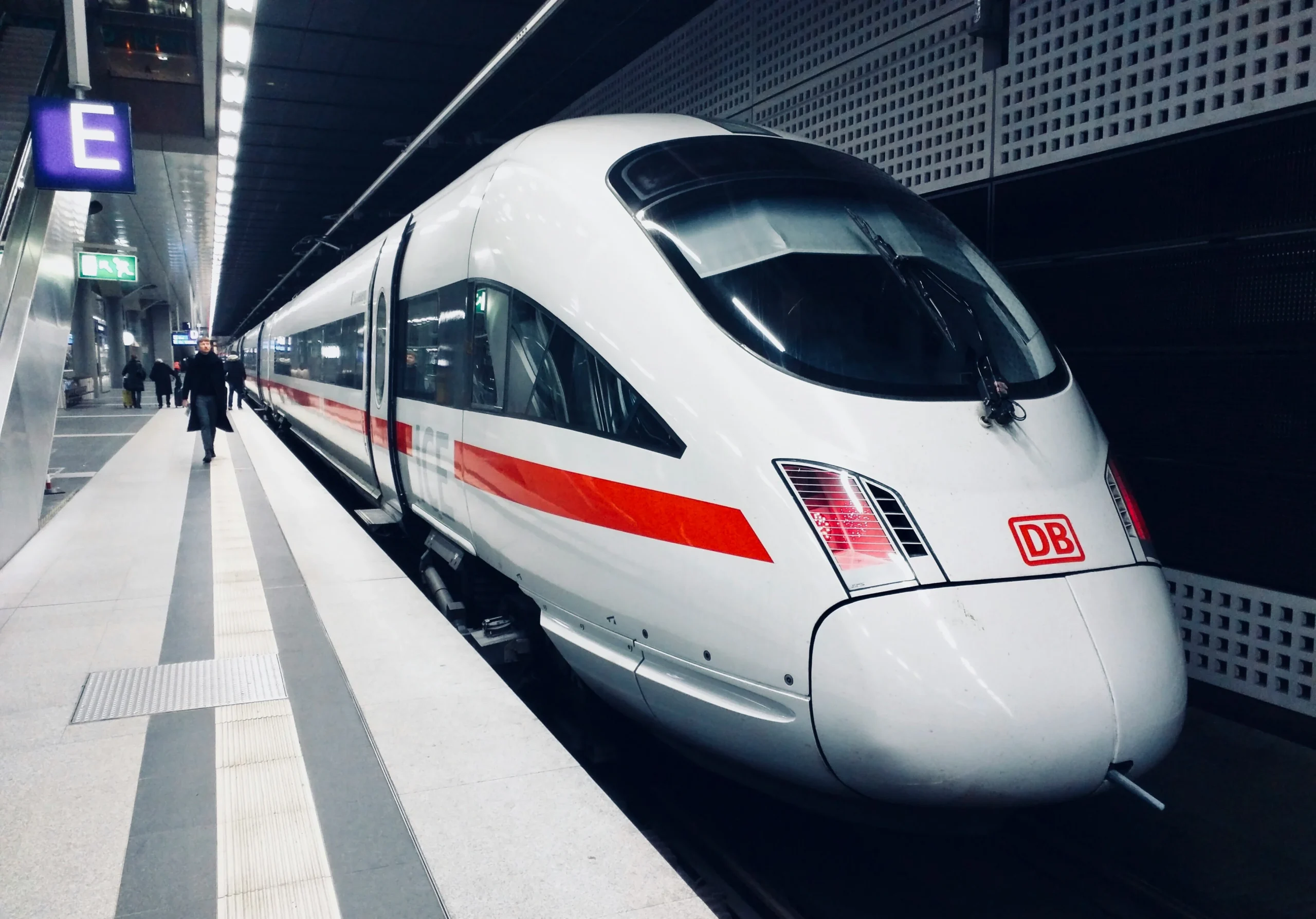 A sleek white high-speed train, a German ICE, with a red stripe and DB logo, is stopped at a modern underground station platform. A person walks away in the distance, and a blue "E" sign is visible.