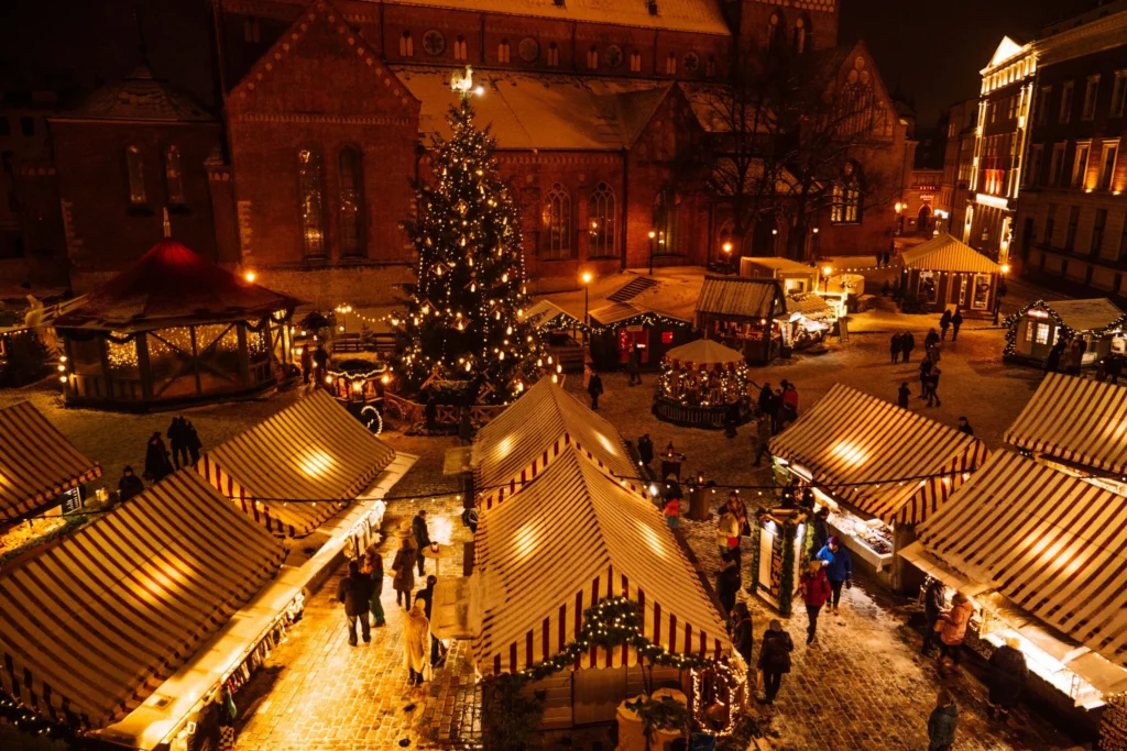 An overhead view of a lively outdoor night Christmas market, with rows of illuminated stalls and a large Christmas tree in the distance, surrounded by people.