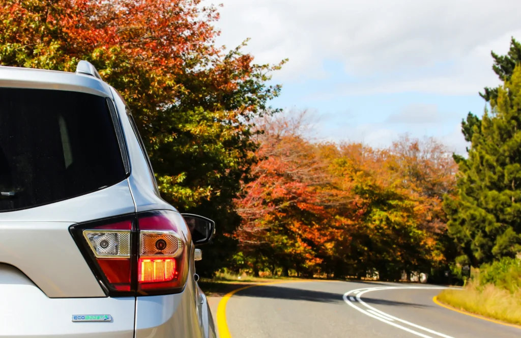 The rear corner of a silver SUV is visible as it sits on a winding road lined with trees displaying vibrant autumn foliage (reds, oranges, and greens) under a partly cloudy sky.