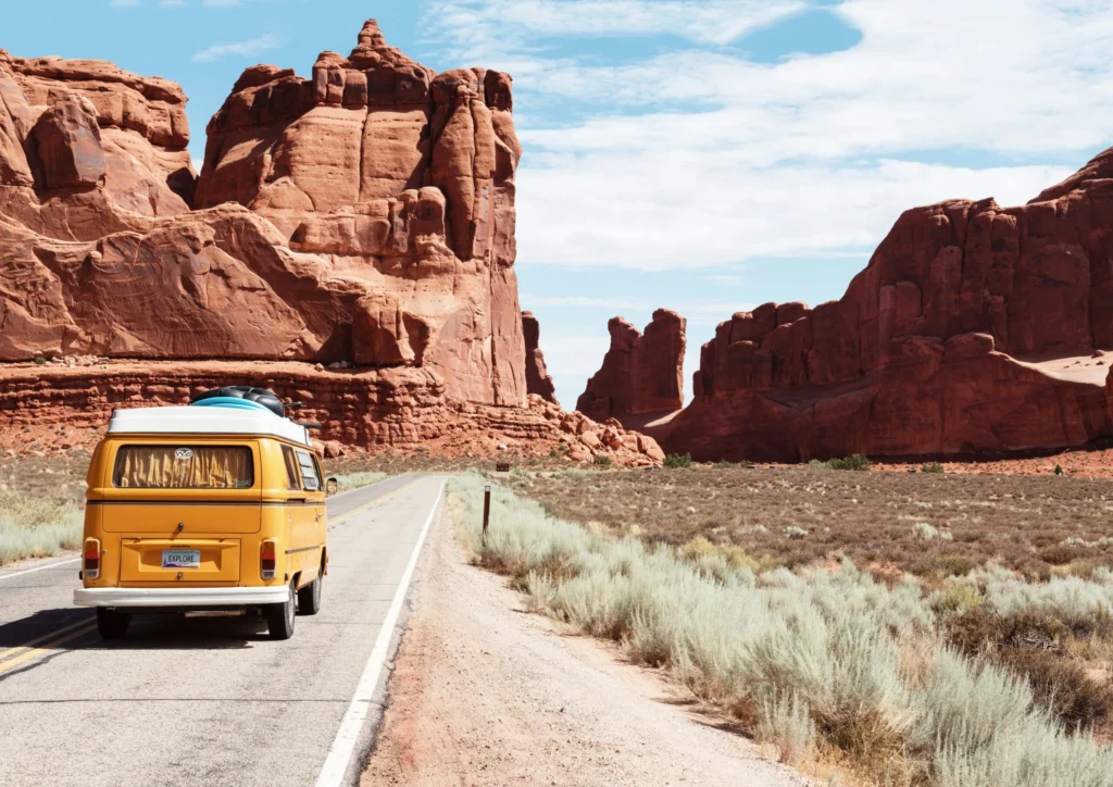 A retro yellow Volkswagen van is parked on the side of a road that stretches through a desert landscape. Towering red rock formations dominate the background under a blue sky with scattered clouds, and dry brush lines the roadside.